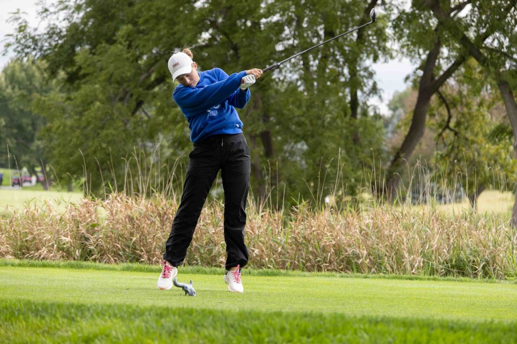 A golfer in a blue sweatshirt and black pants swings a club on a grassy golf course, surrounded by trees and tall grass.