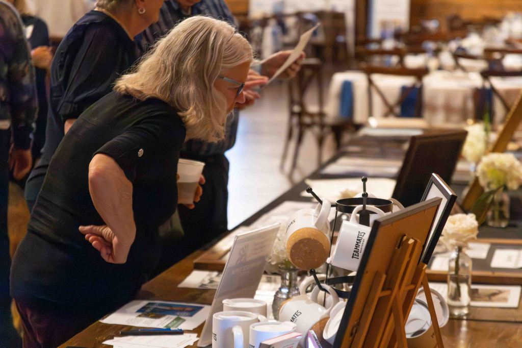 A woman with gray hair and glasses examines materials at a table during an event, holding a cup and standing near other attendees.