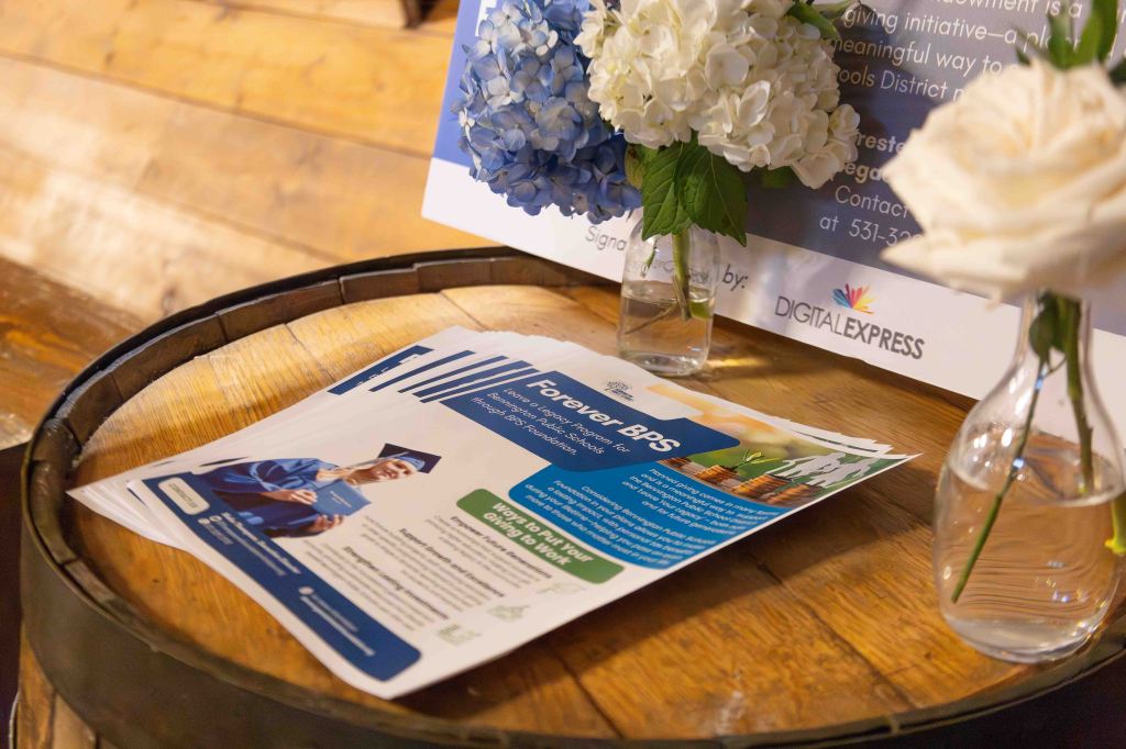 A promotional flyer for the Forever BPS initiative, featuring a graduate in a cap and gown, placed on a wooden barrel. Nearby are two small glass vases with flowers, including white roses and blue hydrangeas.