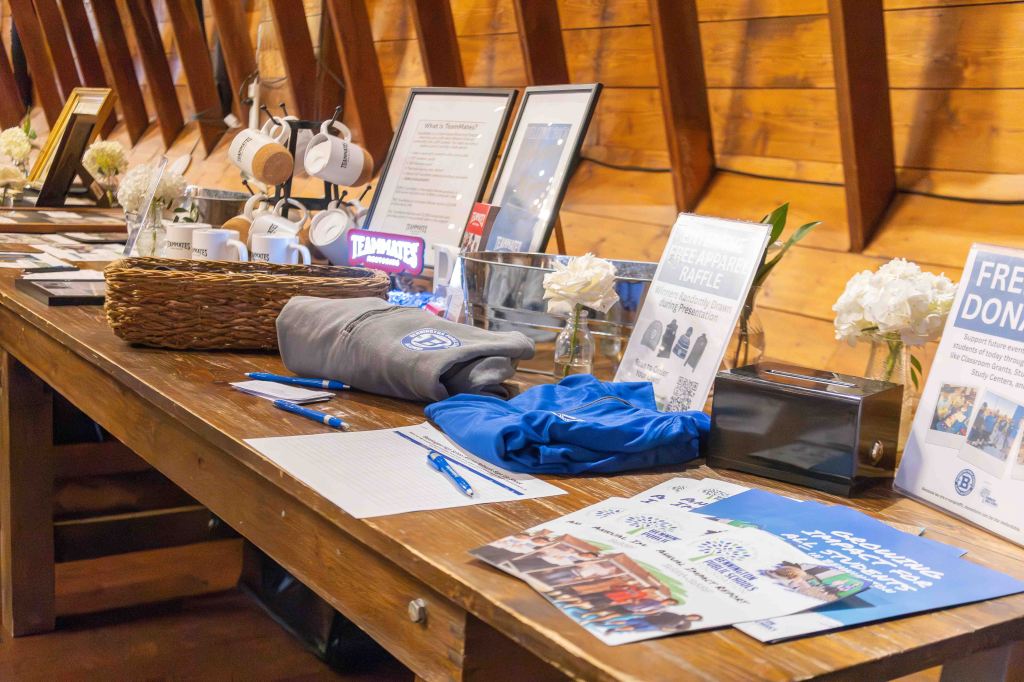 A wooden table displaying various promotional materials, including newsletters, raffle information, and T-shirts, alongside flower arrangements and cups.
