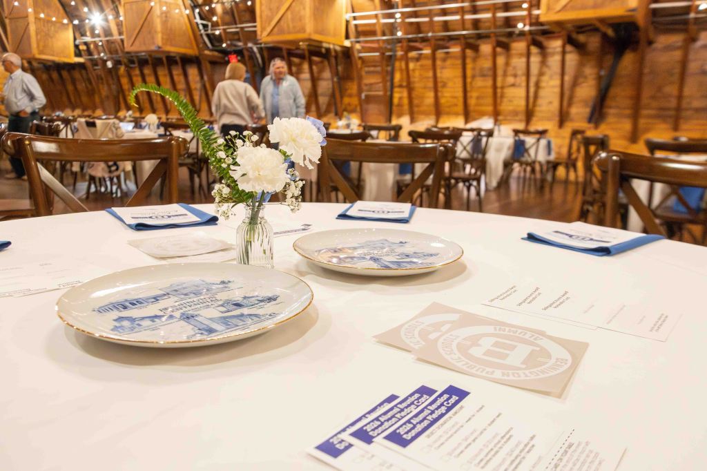 Table setting with white tablecloth, featuring decorative plates, a small vase with flowers, and event menus in a rustic dining space.