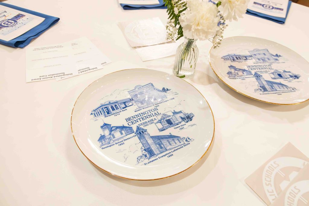 Decorative commemorative plates depicting historical landmarks of Bennington, Nebraska, displayed on a table with floral arrangement and place settings.