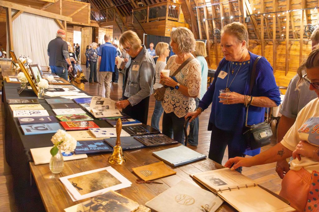 A group of people observing displays of historical documents and photos on a table in a spacious wooden venue.