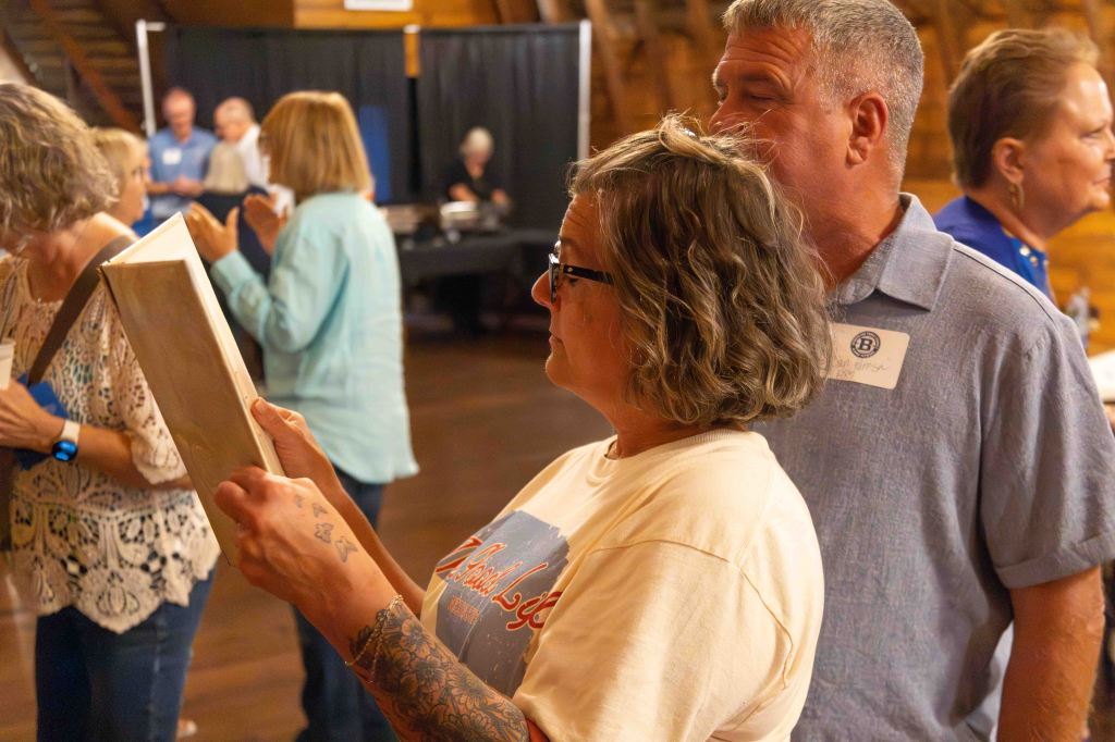 A woman with glasses and curly hair is looking at a book, while a man with short hair stands closely behind her, both engaged in a social gathering with other attendees in the background.