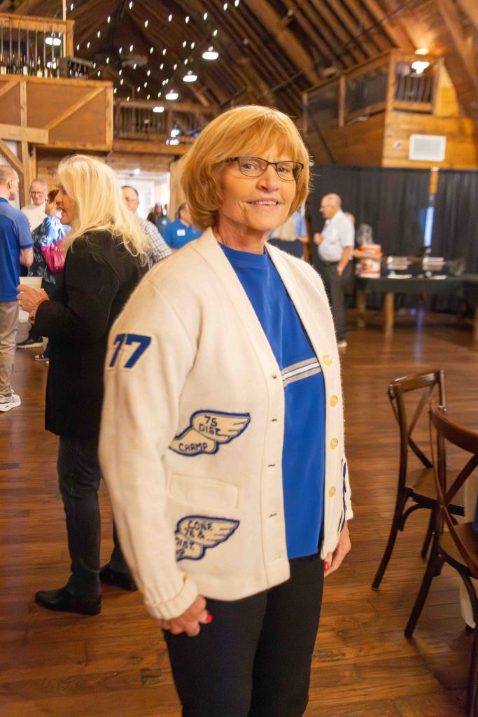 A smiling woman with short blonde hair wearing glasses, a white cardigan with blue details, and a blue shirt, standing in a rustic venue with wooden beams and warm lighting.