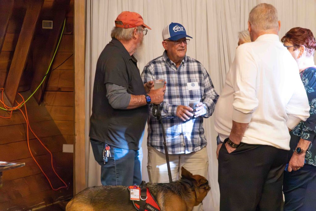 A group of five people socialize indoors, with one individual holding a drink. A dog is also present at their feet.