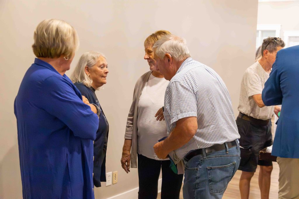 A group of four people engaged in conversation, two older women, one in a dark top and the other in a light sweater, are talking to a man in a striped shirt. Another woman stands nearby, observing the interaction.