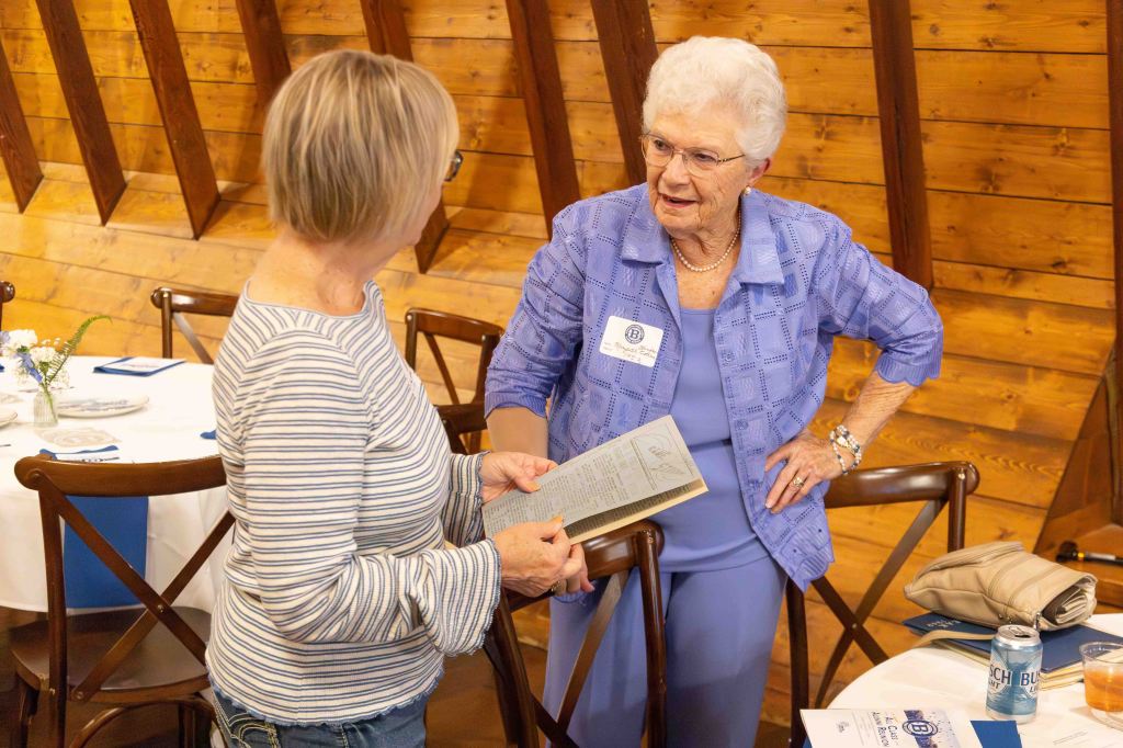 Two women engaged in conversation at a social event inside a warmly lit venue with wooden walls. One woman holds a document while the other listens attentively.