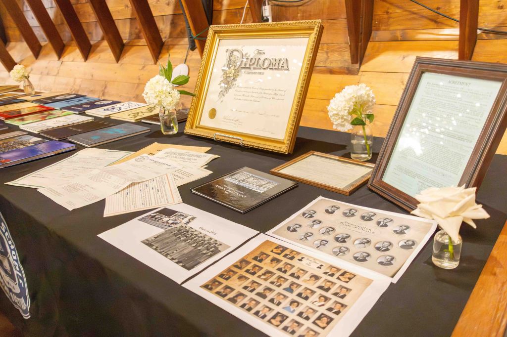 A display table featuring various educational documents, including diplomas, graduation photos, and official papers, with decorative flower arrangements.