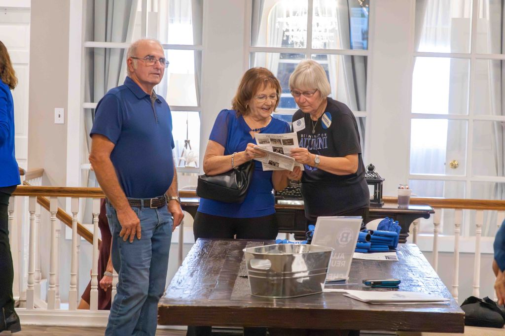 Three individuals are gathered around a table, reviewing a brochure. One man stands with arms crossed, while two women examine the printed material together. The setting is an indoor space with light-colored walls and large windows.