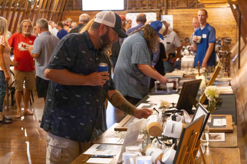 People mingling at a social event inside a wooden venue, with individuals examining items on display tables while holding drinks.