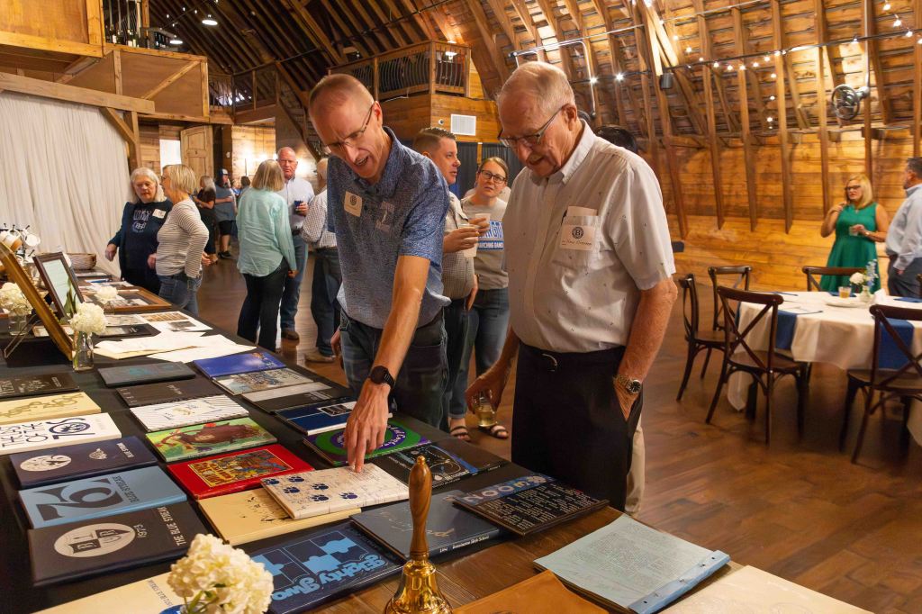 Two men examining a display of colorful booklets and documents on a table in a rustic wooden venue while attendees socialize in the background.