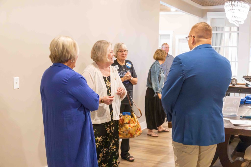 A group of people engaged in conversation in a modern indoor setting. Four women are standing together, two of whom are facing the camera. One woman holds a purse, while another has a friendly expression. In the background, a man and woman are standing further away, with a table displaying materials nearby.