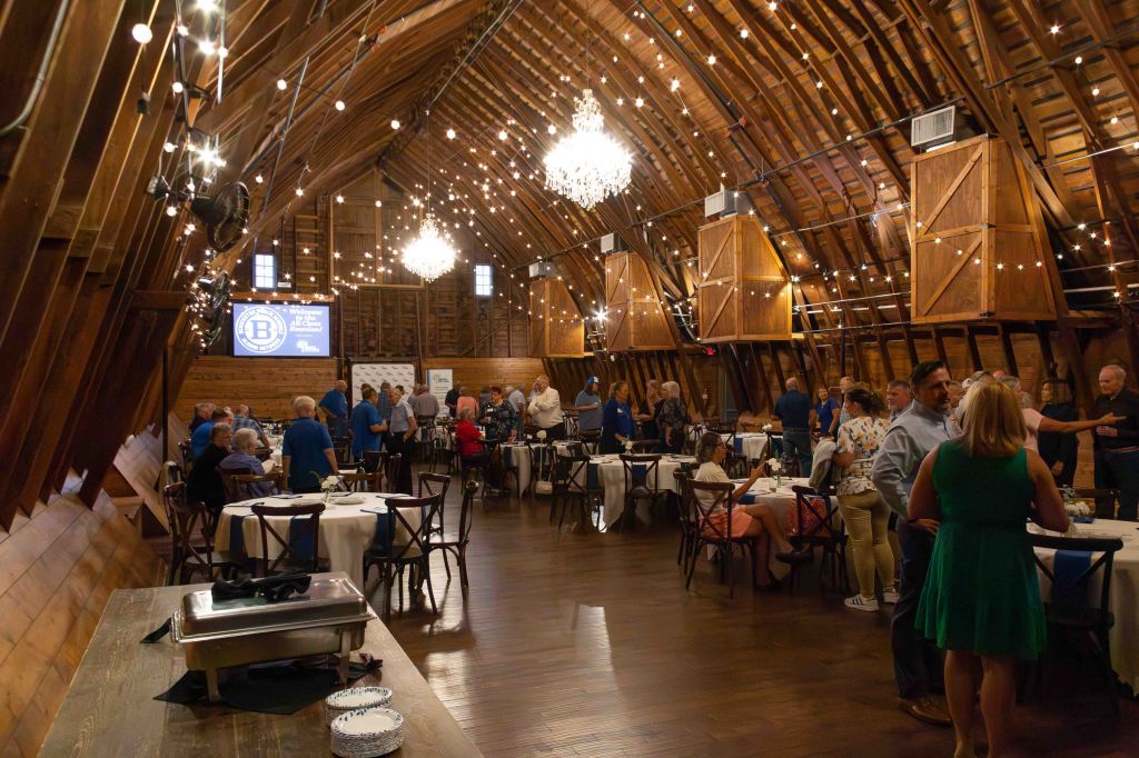 An elegant barn interior with wooden beams and string lights, featuring tables set for dining. Guests in various outfits mingle and socialize in a festive atmosphere.