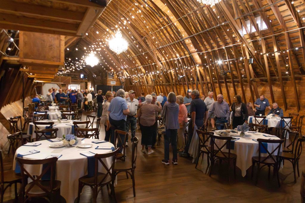 A spacious barn interior decorated for an event, featuring round tables with white tablecloths and blue accents. Guests socialize and mingle in the background under warm ambient lighting, with chandeliers and string lights enhancing the rustic atmosphere.