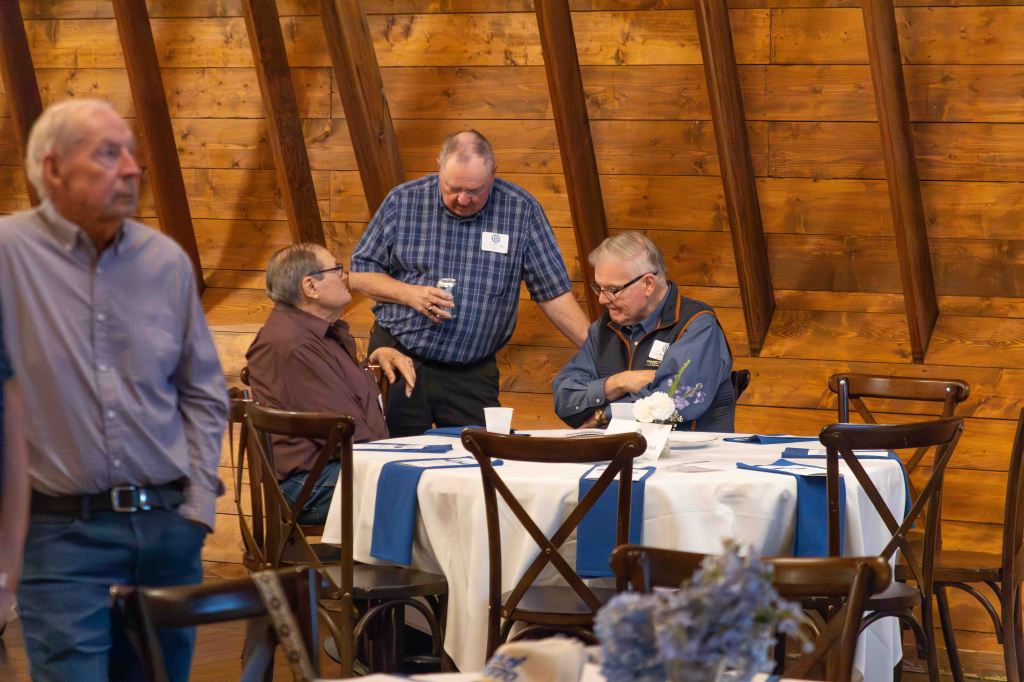 A group of men engaged in conversation at a dining table in a rustic setting with wooden walls.