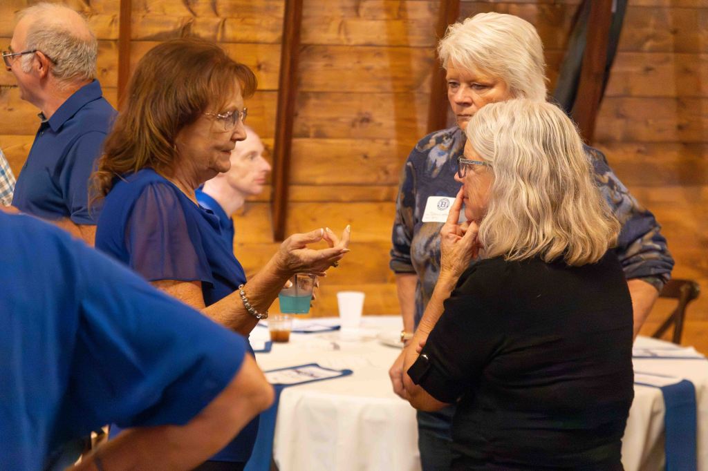 A group of four people engaged in conversation at a social gathering, with a focus on two women discussing while another woman and a man look on. The setting features a rustic wooden background.