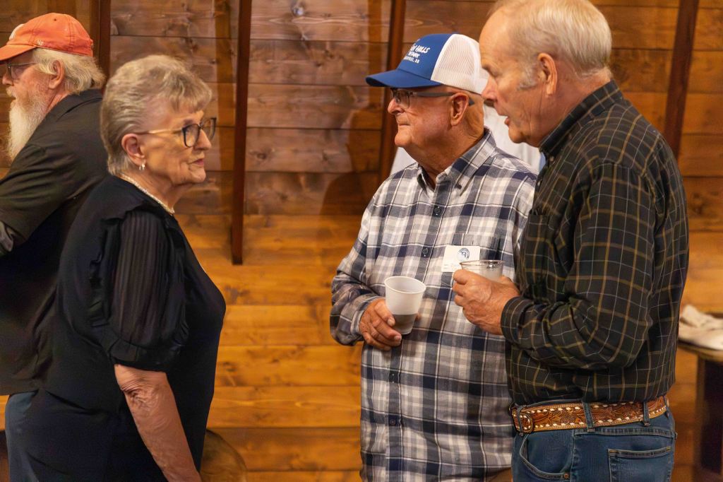 Three individuals engaged in conversation indoors, with a wooden wall in the background. One woman, wearing glasses and a black top, talks to two men: one in a plaid shirt and the other wearing a blue baseball cap and glasses, each holding a cup.