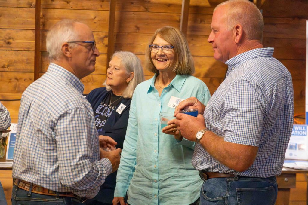 A group of four people engaged in conversation indoors, with wooden walls in the background. Two individuals are smiling and holding drinks, while another is listening attentively. They appear to be enjoying a social gathering.