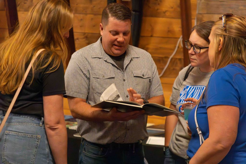 A man in a checkered shirt is showing a book to three women, who are listening and engaging in conversation. The setting appears to be a casual gathering with wooden walls.