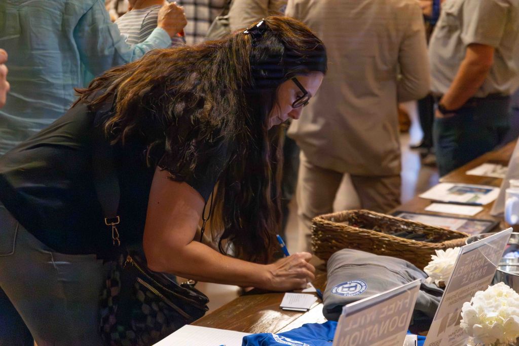 A woman with long dark hair wearing glasses is writing on a notepad while standing at a table with various items and documents. Several people are visible in the background.