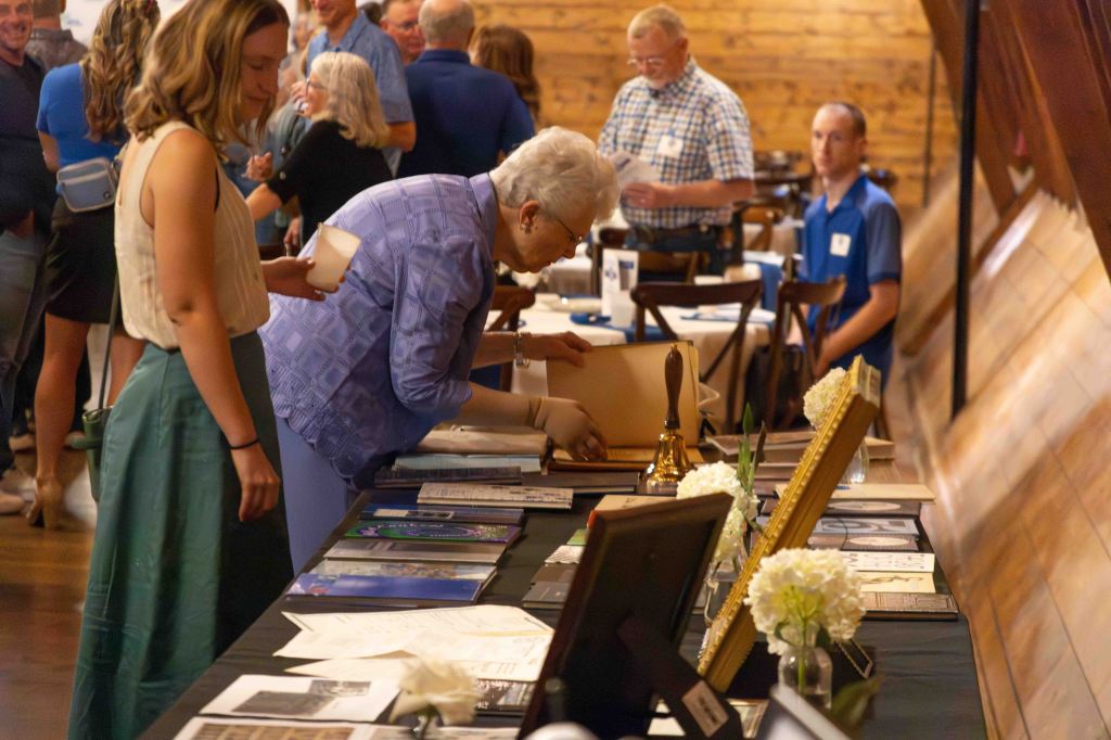 A woman in a blue jacket examines materials on a table at a lively event, while another woman with short hair stands nearby holding a cup. In the background, several attendees engage in conversation.