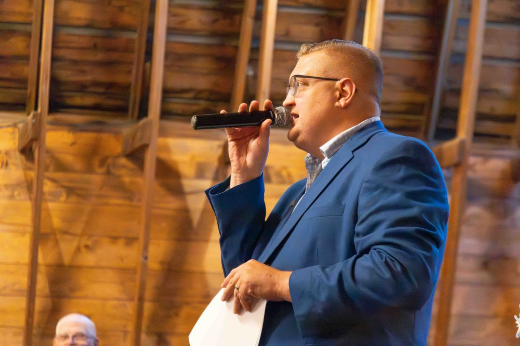 A man in a blue suit presents and holds a microphone while speaking, with wooden barn walls in the background.