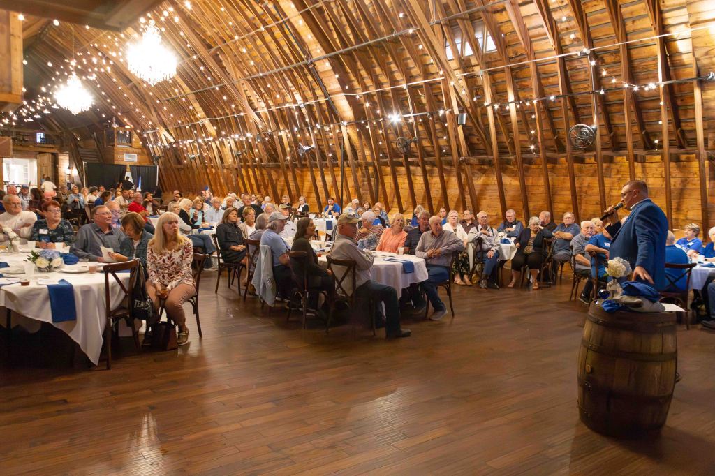 A large indoor gathering in a rustic barn setting, featuring attendees seated at tables with blue and white decorations. A speaker is addressing the audience from a barrel, while chandeliers and string lights hang above.
