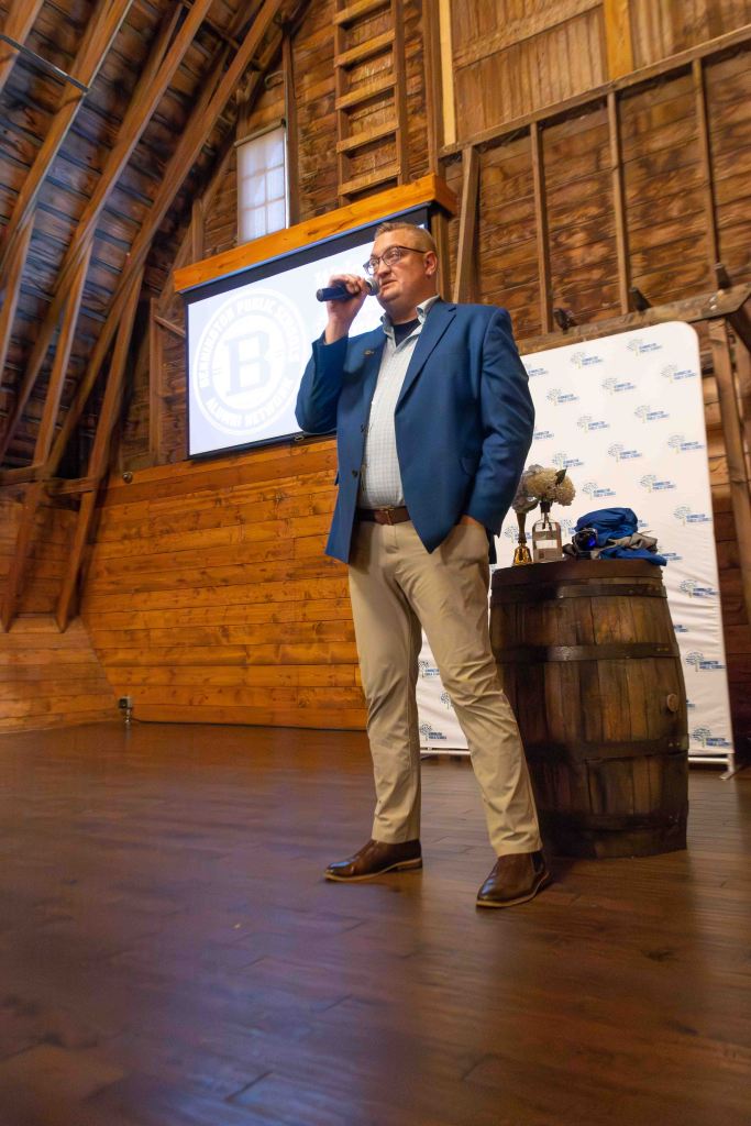 A man standing on a stage inside a wooden barn, holding a microphone and speaking. He is wearing a blue blazer and light-colored pants. A wooden barrel is beside him, and a large screen displays a logo in the background.