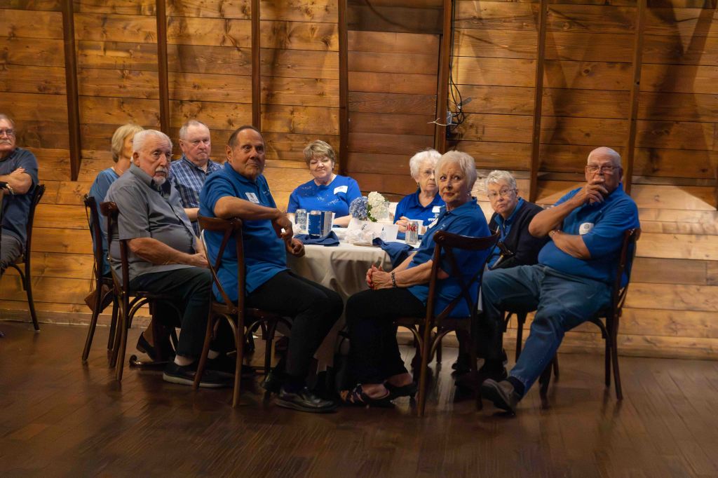 A group of seniors sitting at a table in a cozy venue with wooden walls, wearing blue shirts and looking at the camera.