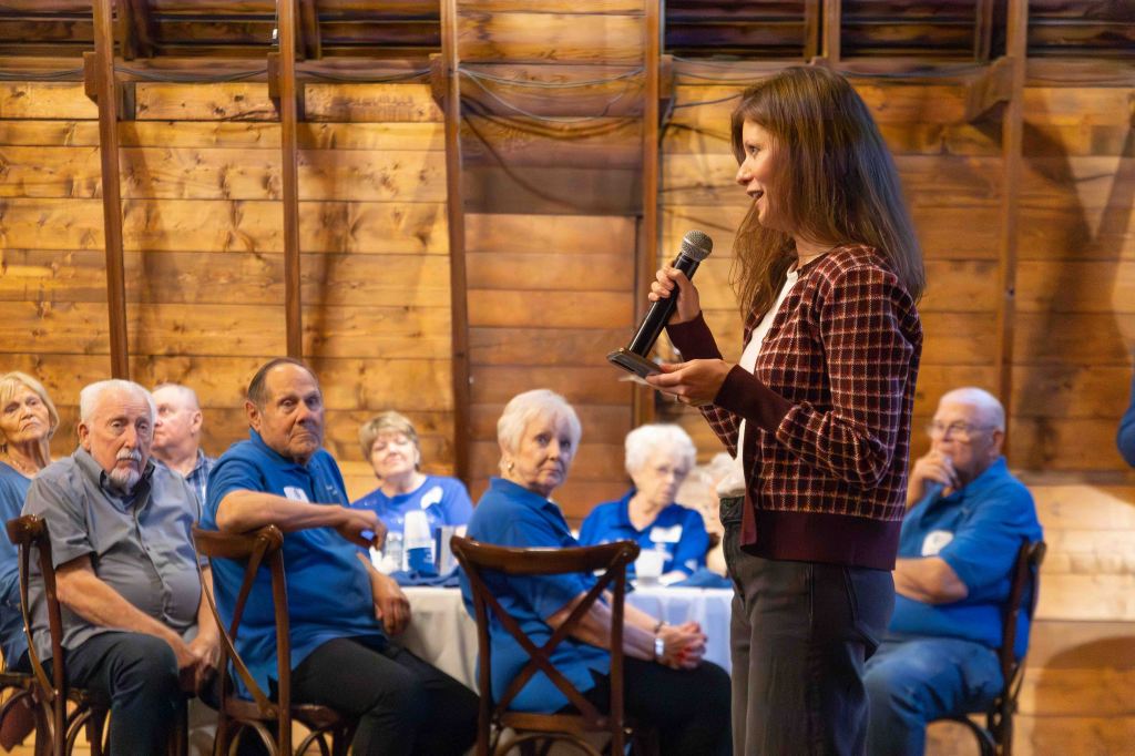 A woman speaking into a microphone in front of an audience of older adults seated at a wooden venue.