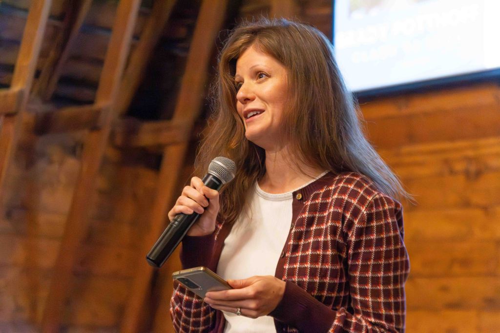 A woman speaking into a microphone while holding a smartphone, wearing a patterned cardigan, in a wooden venue.