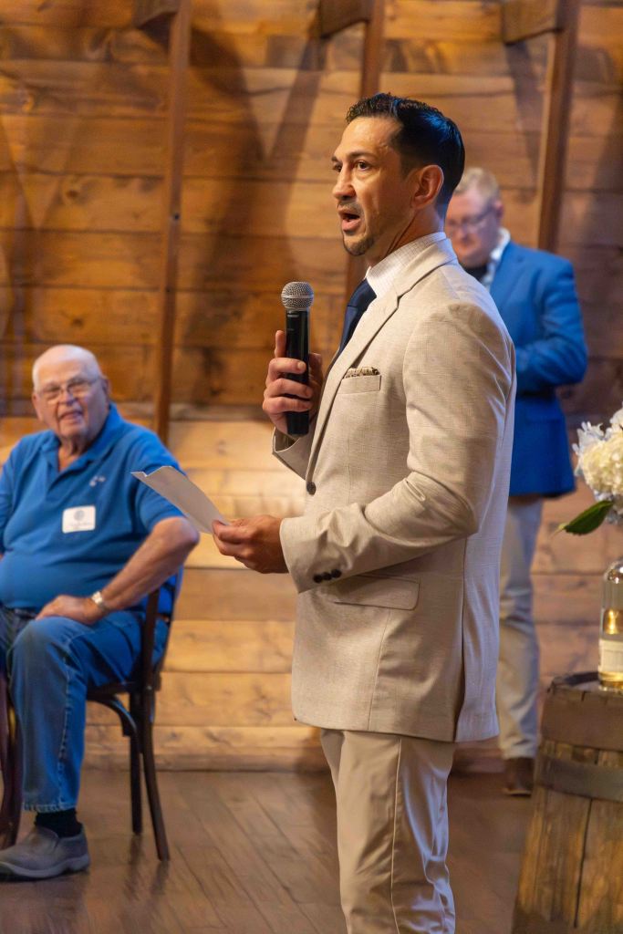 A man in a light-colored suit stands speaking into a microphone while holding a piece of paper, addressing an audience. In the background, an older man in a blue shirt is seated, engaged in the event.