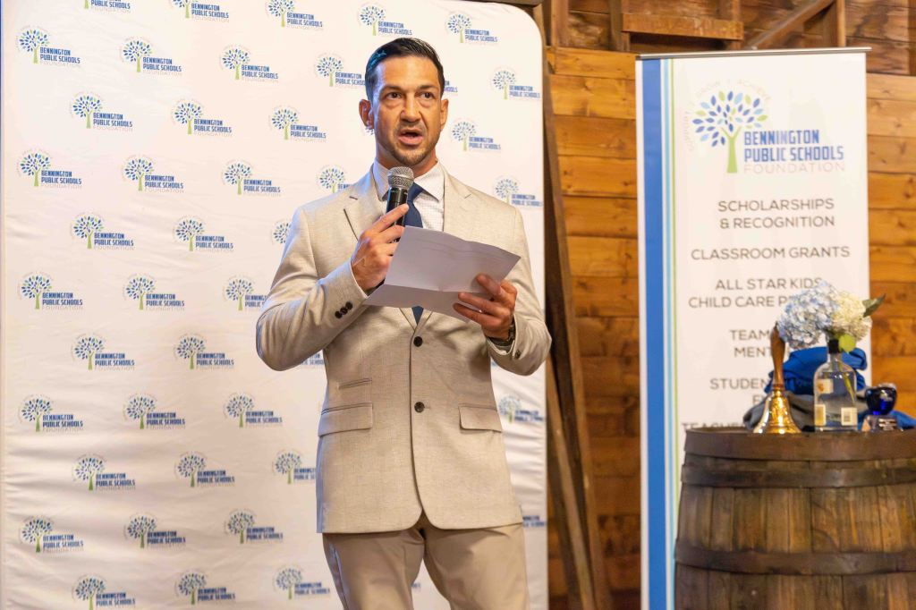 A man in a light-colored suit stands on a stage holding a piece of paper, speaking into a microphone at a Bennington Public Schools Foundation event. The background features a logo and a list of programs supported by the foundation.