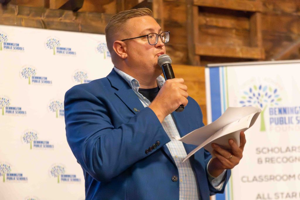 A man in a blue blazer speaking into a microphone while holding notes, in front of a backdrop featuring the Bennington Public Schools logo.