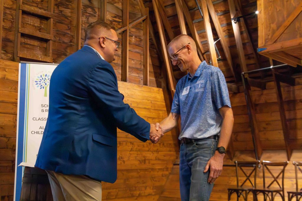 Two men shaking hands in a rustic wooden venue, with a banner in the background indicating a school or community event.
