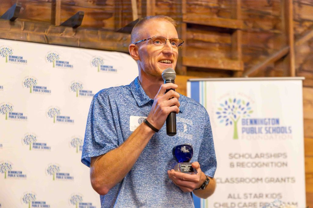 A man in a blue polo shirt holding a microphone and an award, speaking at a Bennington Public Schools Foundation event, with a backdrop featuring the organization's logo.