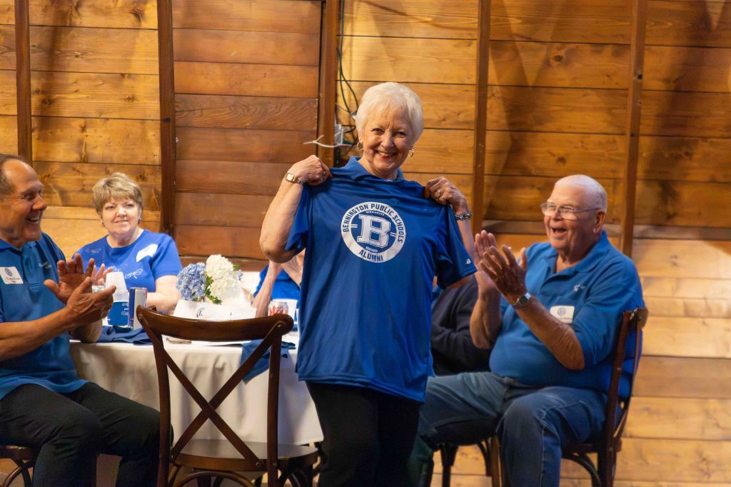 A group of smiling people gathered in a rustic setting, with one woman holding up a blue T-shirt that reads 'B Public Schools Alumni.' Others at the table are cheering and applauding her.