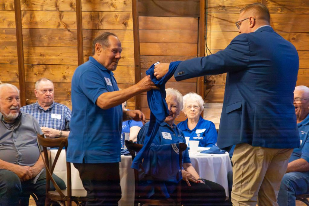 A group of people in blue shirts watching a presentation in a rustic wooden setting. Two men are interacting, with one man presenting a shirt to another seated individual.