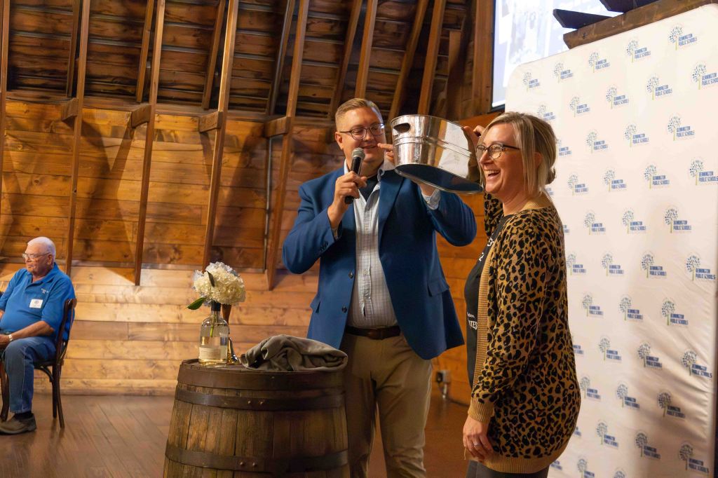A presenter holding a silver bucket while joyfully engaging with a woman who is smiling and wearing a leopard print top, with an audience member seated in the background. The setting features wooden walls and a decorative backdrop.