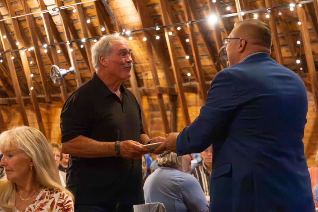 A man in a black shirt is engaged in conversation while receiving a card from another man in a blue suit, inside a rustic barn with wooden beams and soft lighting.