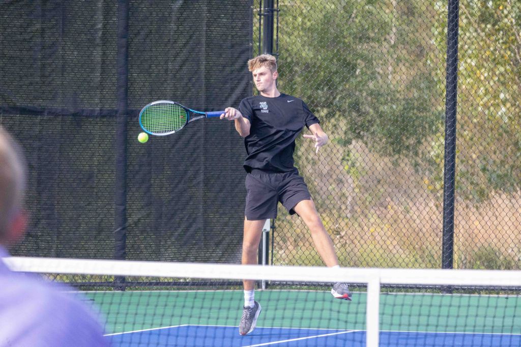 A young male tennis player in mid-air, swinging a racquet to hit a tennis ball during a match, with a court and net in the background.