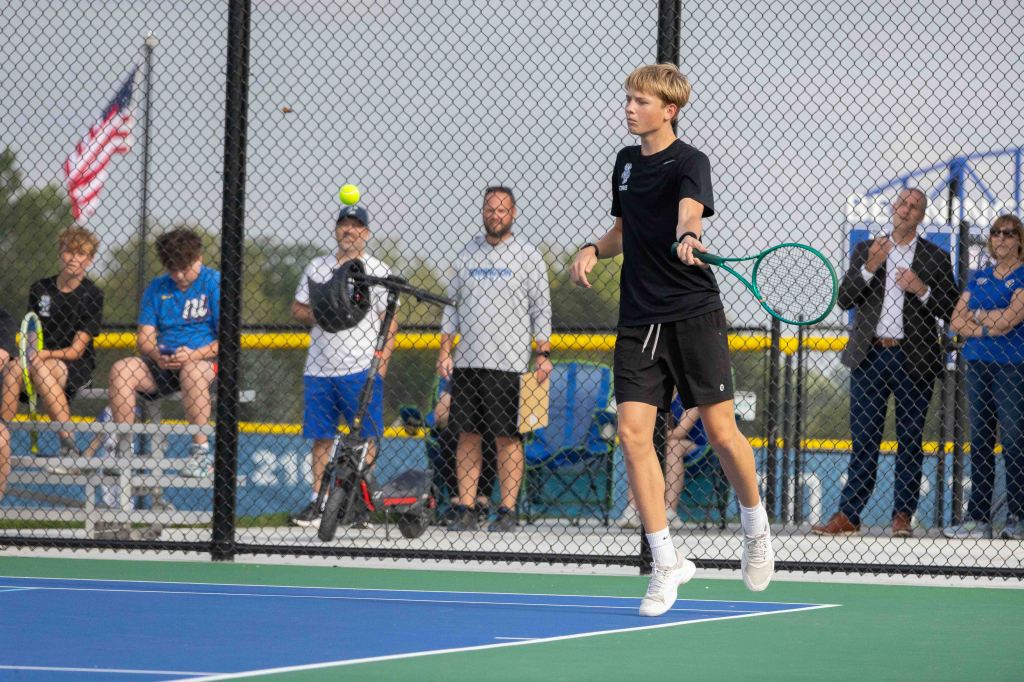 A young male tennis player in a black shirt and shorts prepares to hit a tennis ball on a court, with spectators sitting behind a fence in the background.