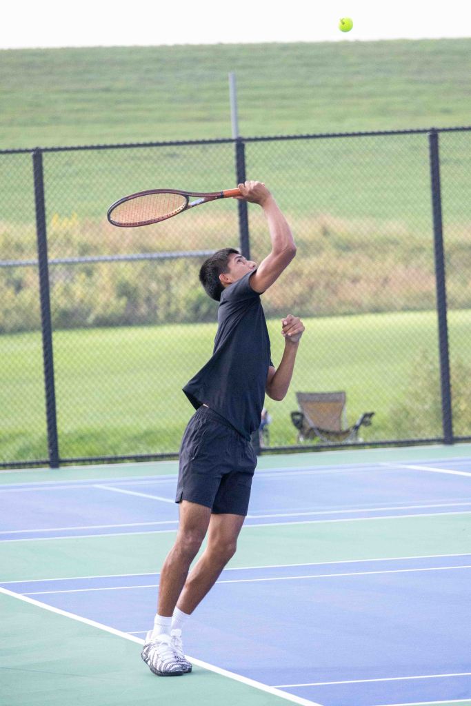 A male tennis player serves the ball on an outdoor court, wearing a black shirt and shorts, with a grassy field in the background.