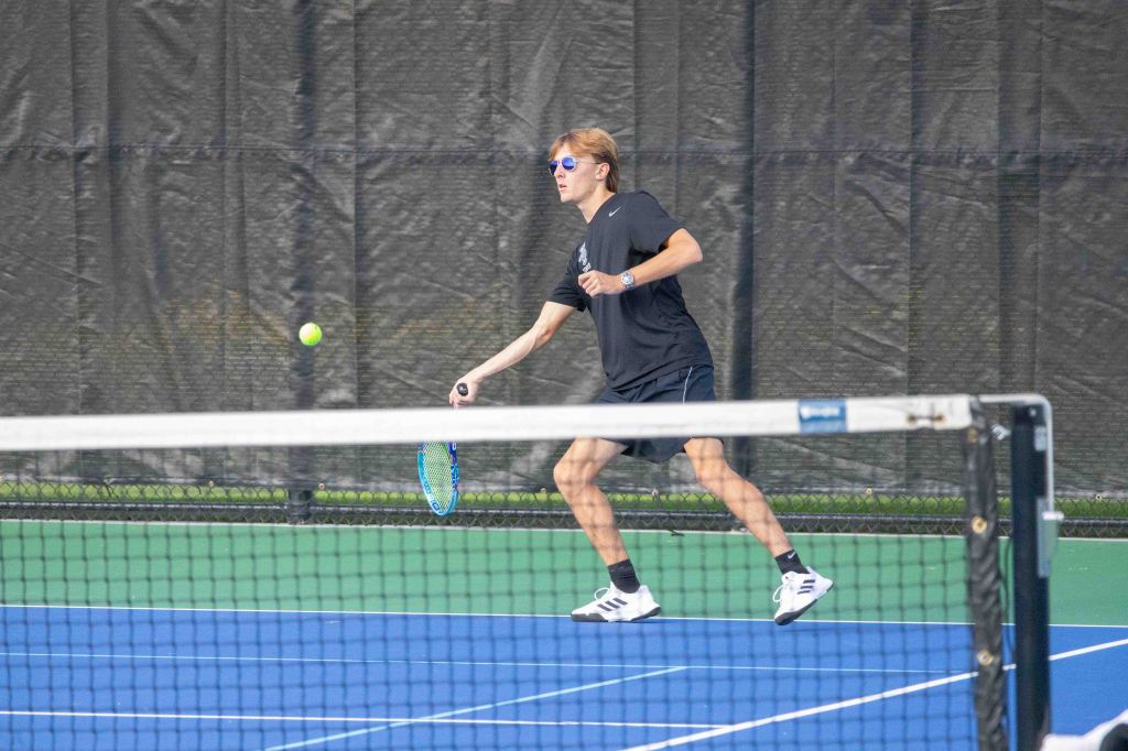 A young male athlete in sunglasses and a black shirt playing tennis, positioned to hit a incoming ball on a blue tennis court.