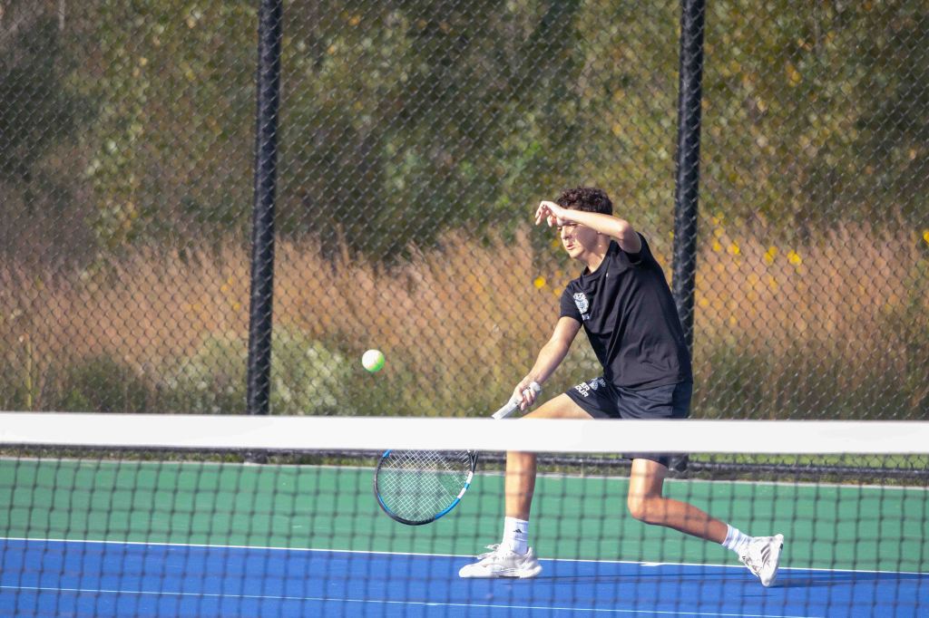 A young male tennis player in a black shirt and shorts is executing a backhand shot while hitting a tennis ball on a bright blue court.