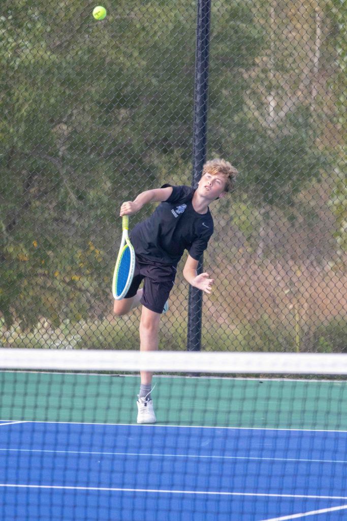 A young male tennis player serving the ball on a blue tennis court, wearing a black shirt and shorts, with a focused expression.