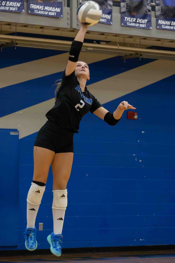 A volleyball player jumps to set the ball during a game, wearing a black jersey with the number 2, black shorts, and white knee pads in a gym with blue walls.