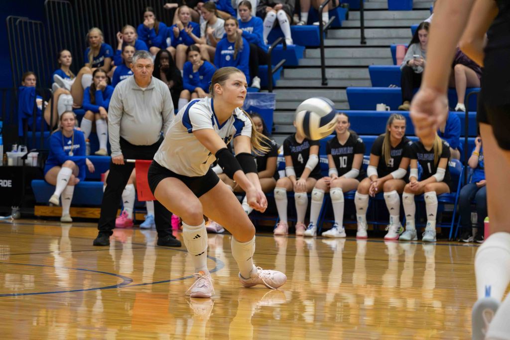 A volleyball player in a white jersey is diving to receive the ball during a match, with teammates and a coach in the background watching attentively.