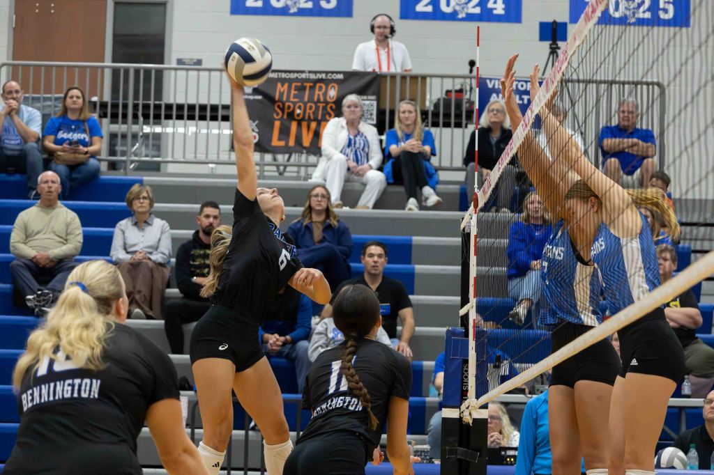 A volleyball player jumps to spike the ball over a net during a match, with spectators watching in the background.